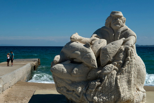 Το άγαλμα του ψαρά που βρίσκεται στην παραλία της Ιερισσού με φόντο τη γαλάζια θάλασσα και τον γαλάζιο ουρανό.