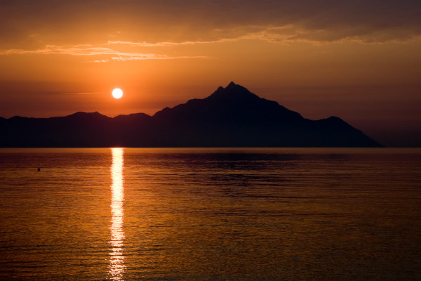 Η ανατολή του ηλίου πάνω από τα βράχια του Αγίου Όρους ενώ ο ήλιος αντανακλάται στα ήρεμα νερά του Αιγαίου πελάγους.