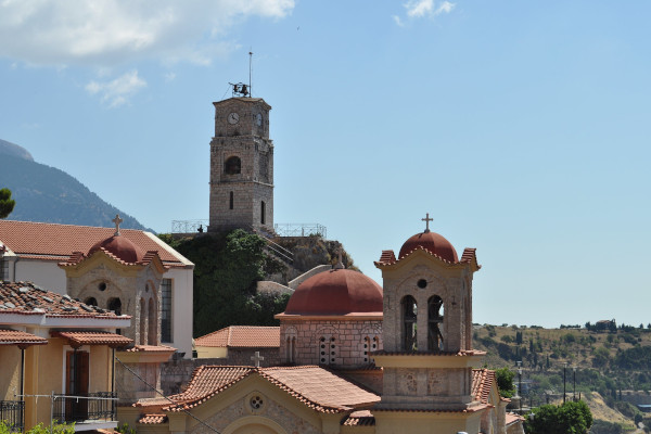 Μια εικόνα που δείχνει τον Πύργο του Ρολογιού της Αράχωβας ανάμεσα στα σπίτια και την εκκλησία του οικισμού.