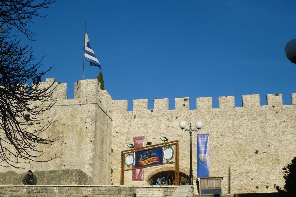 Ένας πέτρινος τοίχος και μια από τις πύλες του Κάστρου της Άρτας κάτω από μια ελληνική σημαία.