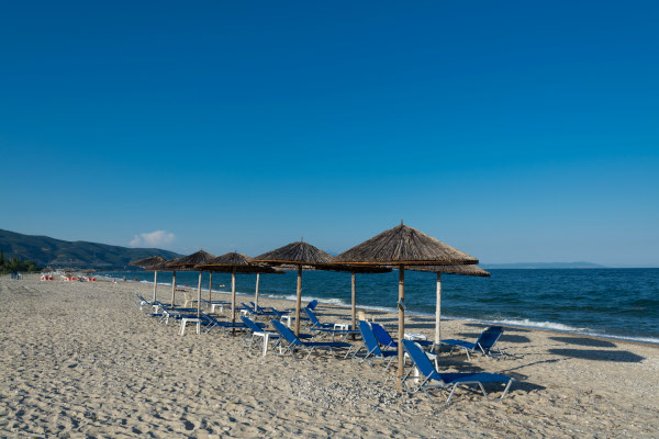 Ομπρέλες και ξαπλώστρες στην παραλία της Ασπροβάλτας κάτω από έναν γαλάζιο ουρανό.