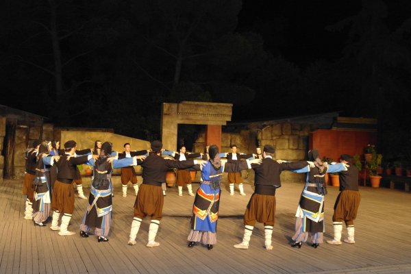 Γυναίκες και άνδρες με παραδοσιακή ενδυμασία χορεύουν κυκλικά σε μια μεγάλη σκηνή. 