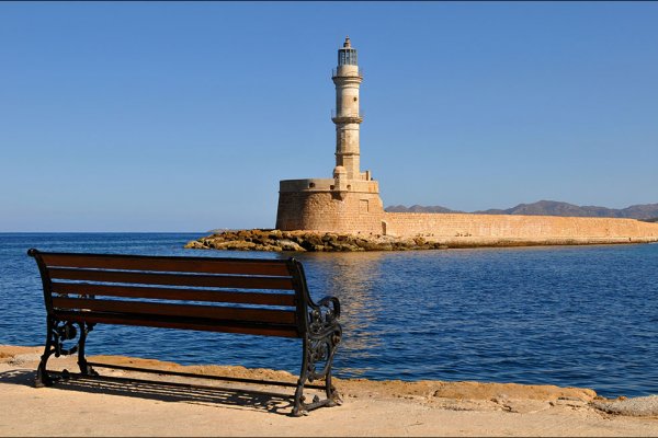 Ένα παγκάκι δίπλα στην καταγάλανη θάλασσα με θέα τον Φάρο των Χανίων που είναι χτισμένος με ανοιχτόχρωμες πέτρες.