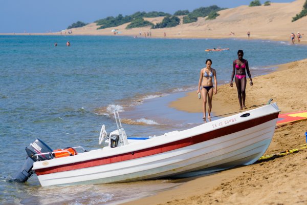 Δύο γυναίκες περπατούν μπροστά από μια βάρκα με φόντο τους αμμόλοφους στην παραλία Ίσσος, Αγ. Γεώργιος.