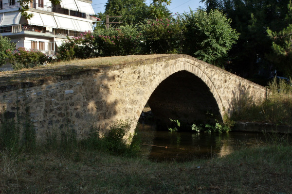 Το αρχαίο πέτρινο γεφύρι του Κιουπρί πάνω από ένα ρυάκι περιτριγυρισμένο από δέντρα.
