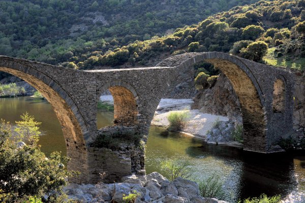 Το τοξωτό πέτρινο γεφύρι πάνω από τον ποταμό Κομψάτο και στο βάθος λόφοι καλυμμένη από θάμνους.