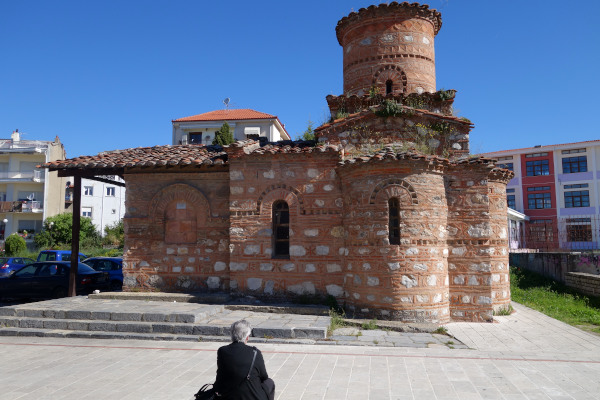 Μια φωτογραφία της βυζαντινής εκκλησίας της Παναγίας της Κουμπελιδικής στην Καστοριά με μια ηλικιωμένη γυναίκα να την κοιτάζει.