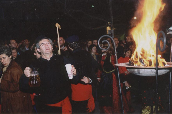 Άνθρωποι με παραδοσιακά ρούχα, κρατώντας μια κανάτα κρασί, τραγουδούν γύρω από μια φωτιά σε μεταλλικό σκεύος.