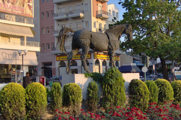 Το άγαλμα του Βουκεφάλα περιτριγυρισμένο από θάμνους και λουλούδια, με φόντο μερικές πολυκατοικίες της Λάρισας.