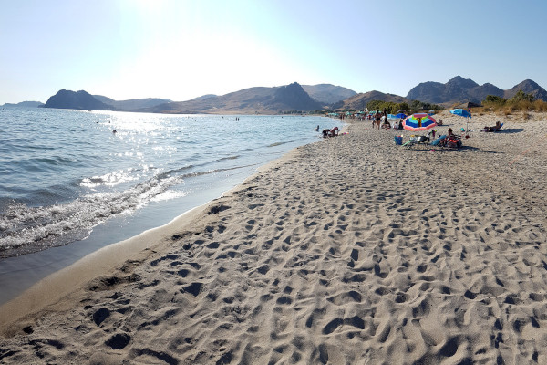 Φωτογραφία της λευκής άμμου, της θάλασσας, μερικών τουριστών και λόφων στο βάθος στην παραλία Ευγάτης στη Λήμνο.