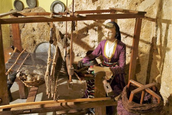 Αναπαράσταση μιας γυναίκας που εργάζεται σε έναν παλιό ξύλινο αργαλειό σε μια εικόνα όπου κυριαρχούν τα χρώματα της γης.