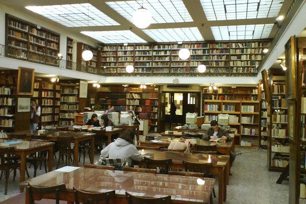 Άνθρωποι που διαβάζουν σε τραπέζια ανάμεσα σε τοίχους από ράφια κάτω από φυσικό φως στη Δημοτική Βιβλιοθήκη της Πάτρας.