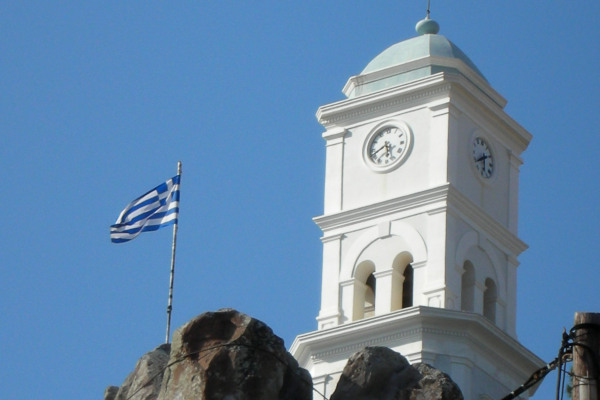 Μια ελληνική σημαία δίπλα στον λευκό ρολόι του Πόρου και ένας γαλάζιος ουρανός φόντο.