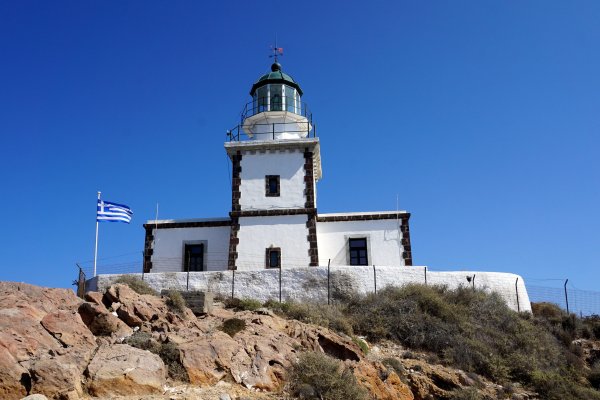 Ένας λευκός φάρος με καφέ γωνίες που σχηματίζουν τετράγωνα βρίσκεται στην κορυφή ενός βραχώδους λόφου, και μια ελληνική σημαία.