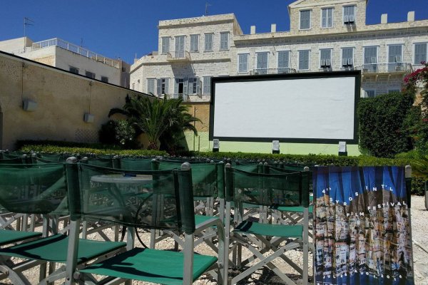 Πράσινες καρέκλες σκηνοθέτη μπροστά από μια μεγάλη οθόνη με λευκά κτίρια στο βάθος.