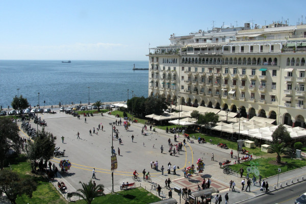 Μια πανοραμική εικόνα που δείχνει ανθρώπους στην πλατεία Αριστοτέλους με φόντο ένα λευκό κτίριο και θέα τη θάλασσα.