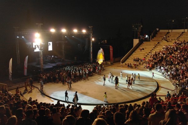 Ένα μισογεμάτο αμφιθέατρο με χορωδία και παιδιά σε μια φωτισμένη σκηνή κατά τη διάρκεια της νύχτας.