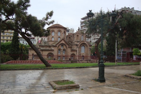 Η πλινθόκτιστη βυζαντινή εκκλησία πίσω από το πλακόστρωτο και λίγα δέντρα  περιβάλλεται από πολυκατοικίες.