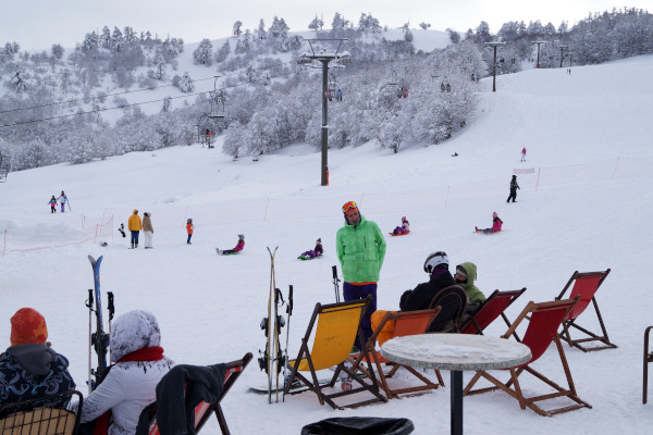 Άνθρωποι με χειμωνιάτικα ρούχα κάθονται σε καρέκλες δίπλα στον εξοπλισμό σκι τους με φόντο την πλαγιά της Βασιλίτσας.